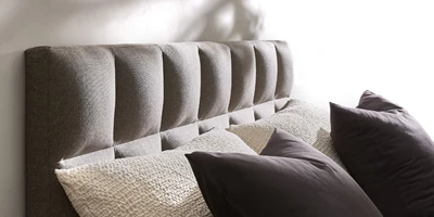 Design uden navn - 2025-03-13T134744.809.jpg Säng med stoppad gavel med breda, lodräta paneler i grått tyg och stora kuddar.