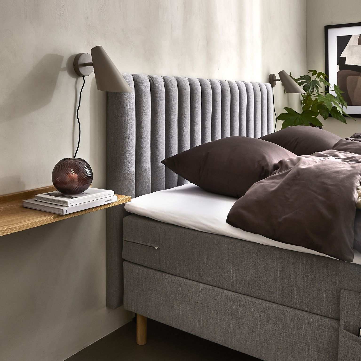 Modern säng med grå, vertikalt stoppad sänggavel, mörka sängkläder och vägghylla i trä