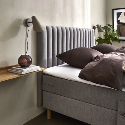 Modern säng med grå, vertikalt stoppad sänggavel, mörka sängkläder och vägghylla i trä