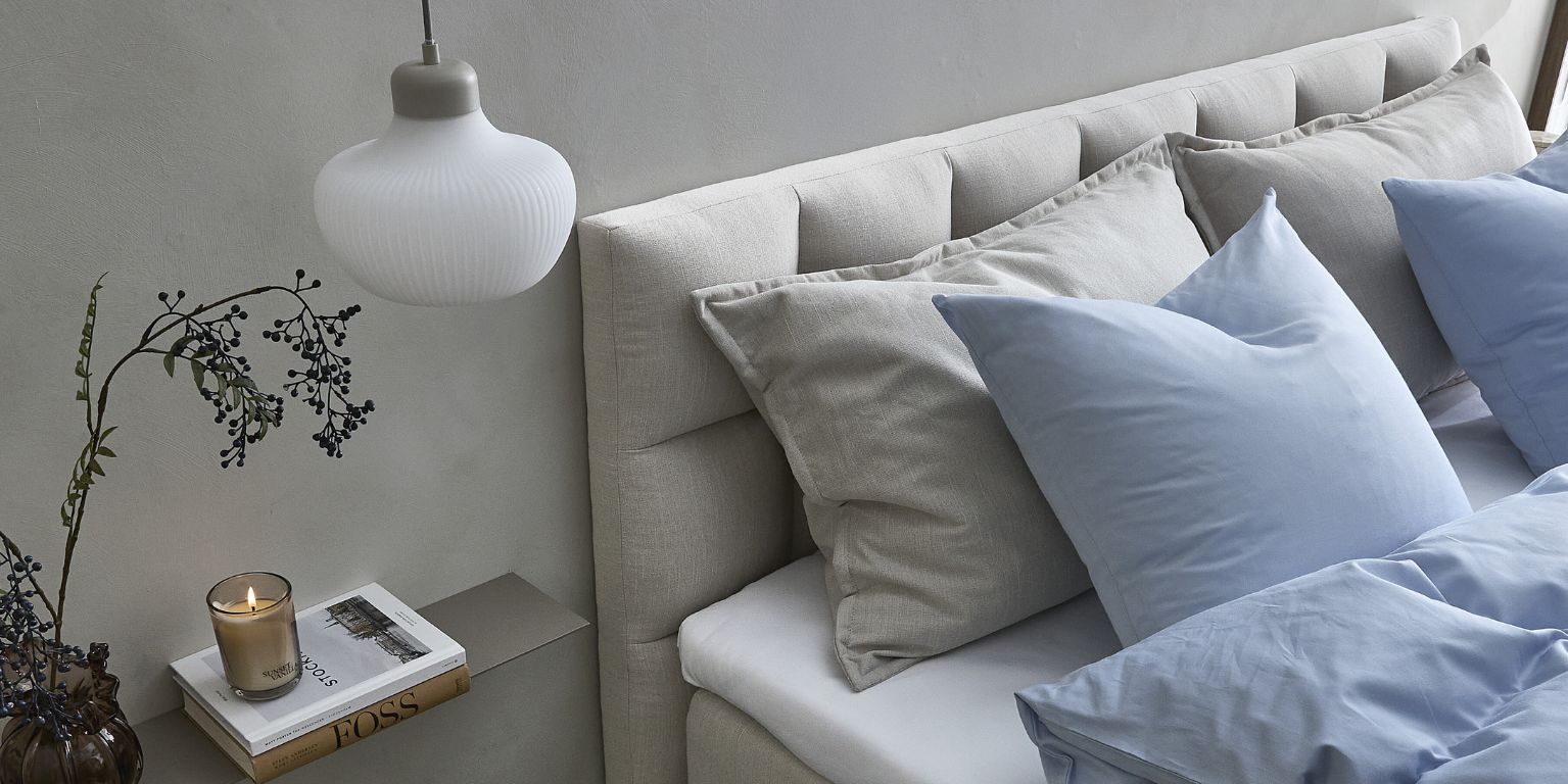 Säng med ljus, horisontellt stoppad gavel, blåa sängkläder, levande ljus och liten vägghylla.