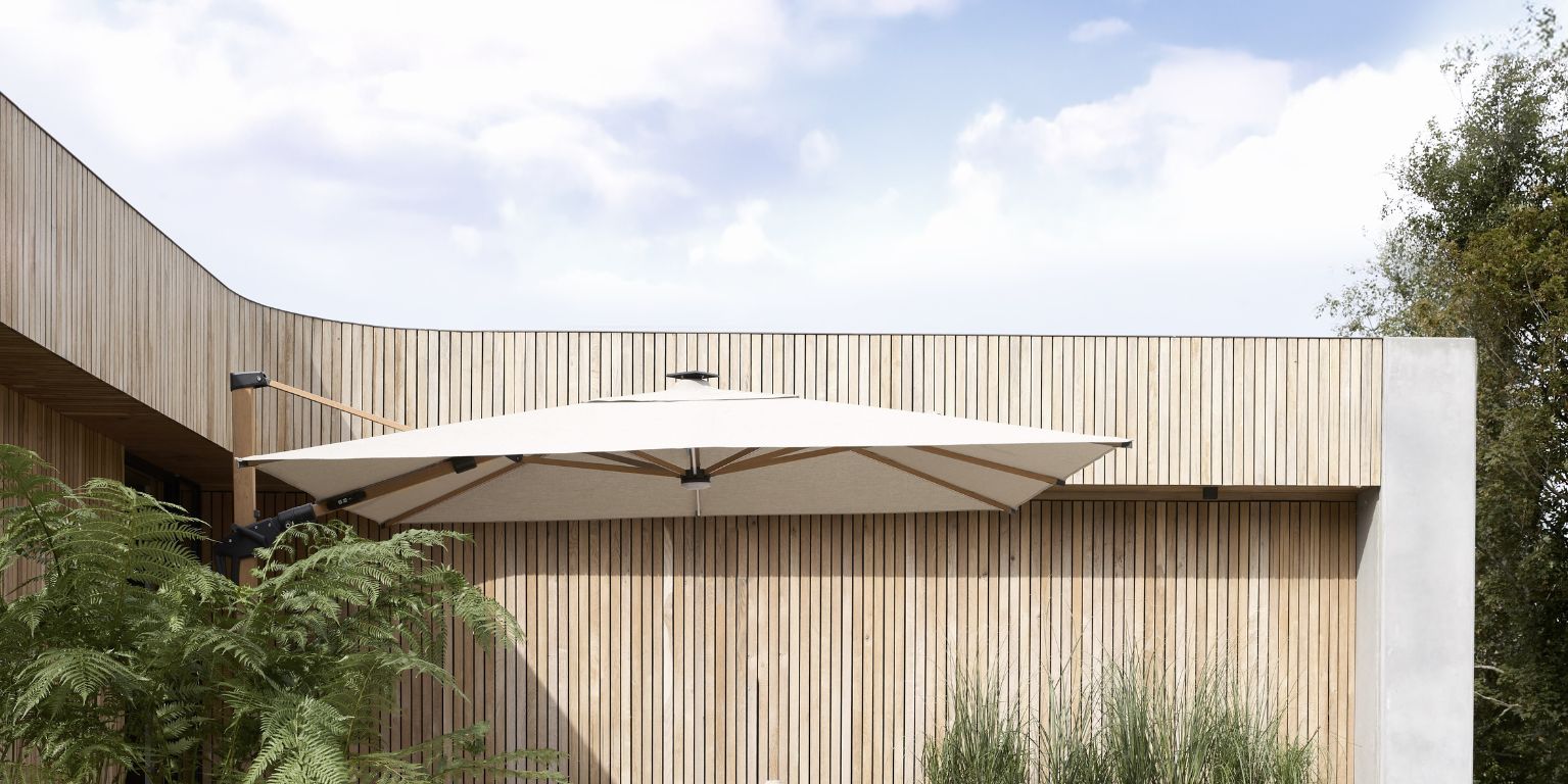 Stor fyrkantig parasoll monterad på en modern byggnad med vertikala träpaneler och frodiga gröna växter i förgrunden