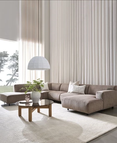 Ljust vardagsrum med en beige u-soffa, soffbord med glasskiva och vita gardiner i bakgrunden