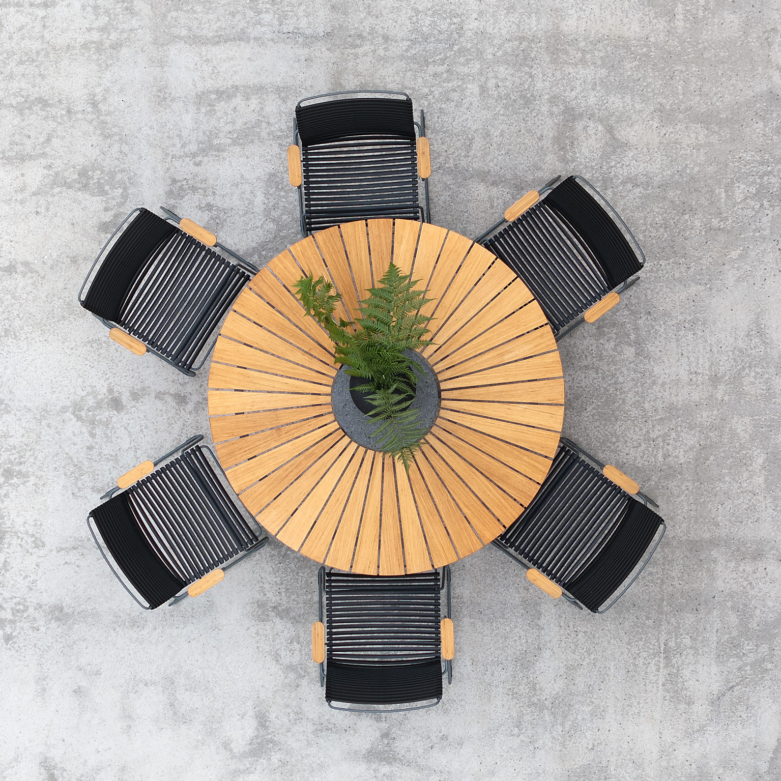 Bild tagen ovanifrån på ett runt utebord i teakträ, omgivet av sex stycken stolar med svarta plastlameller och trähandtag.