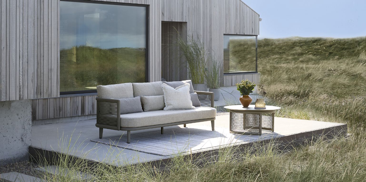 Utomhusterrass i sanddynerna vid trähus med loungesoffa och bord.
