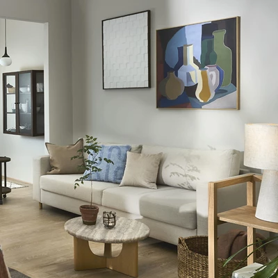 Ljus bäddsoffa i vardagsrum med soffbord, vägghängd hylla och bilder på väggen.