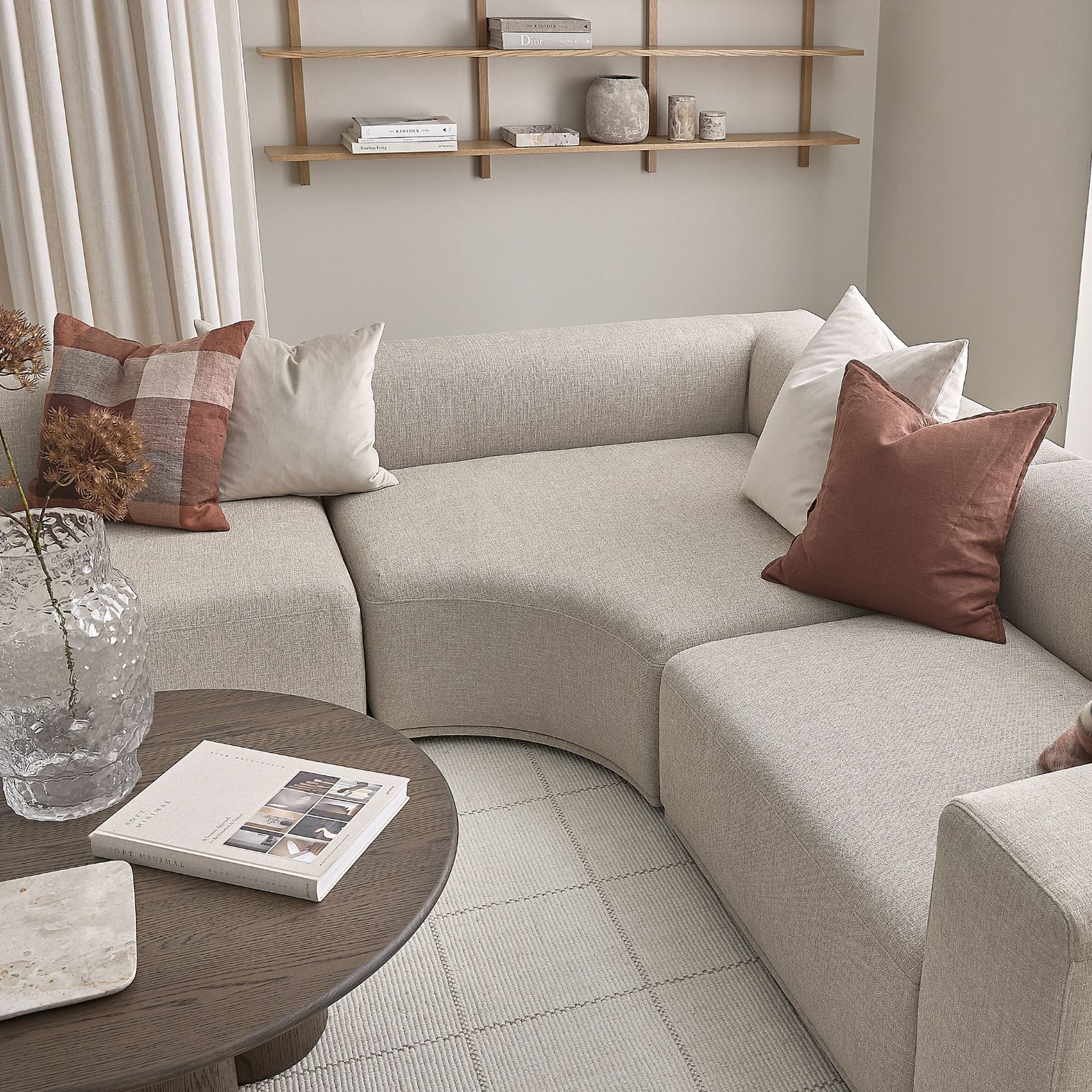 Hörnsoffa i ljusgrått med böjt hörnmodul stående på rutigt golv. Framför står ett soffbord i mörkt trä.