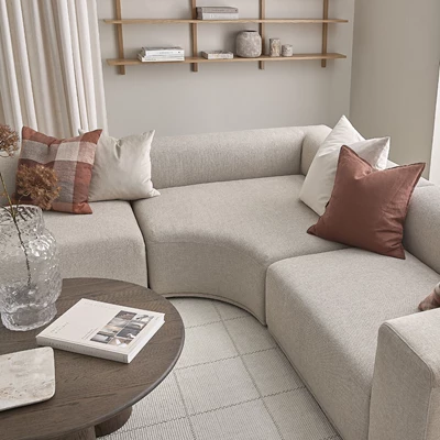 Hörnsoffa i ljusgrått med böjt hörnmodul stående på rutigt golv. Framför står ett soffbord i mörkt trä.