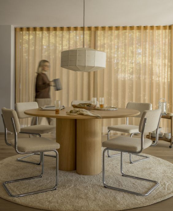 Vardagsrum i varma färger med ett runt matbord och stoppade grå stolar i centrum.