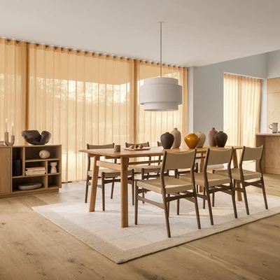 Matbord i trä med sex stolar i en ljus matsal med stora fönster och beiga gardiner.