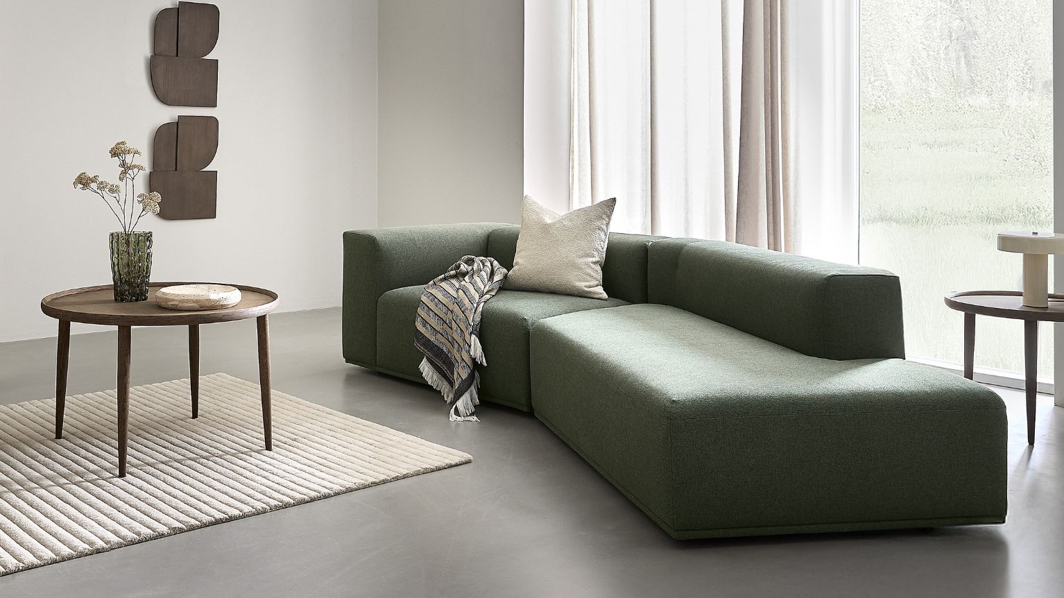 Grön hörnsoffa med kudde och filt i ett ljust vardagsrum med runt träbord och matta.