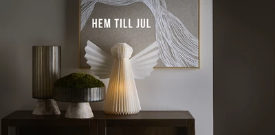 Julig dekoration med lysande pappersängel, vas och skål med mossa framför tavla med texten “Hem till jul”.